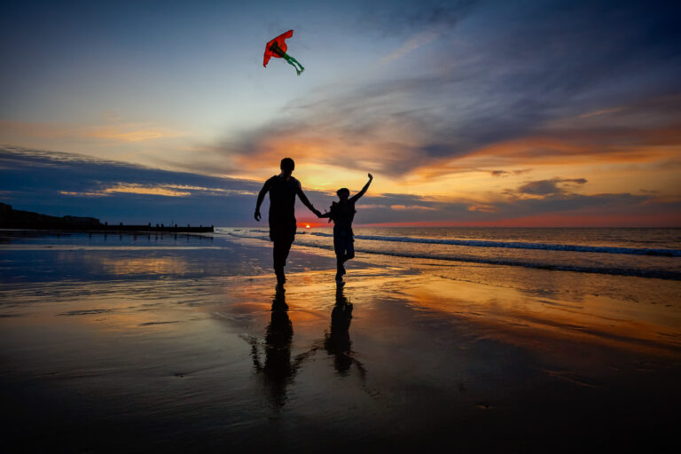 A young couple together flying a kite at sunset on the beach.